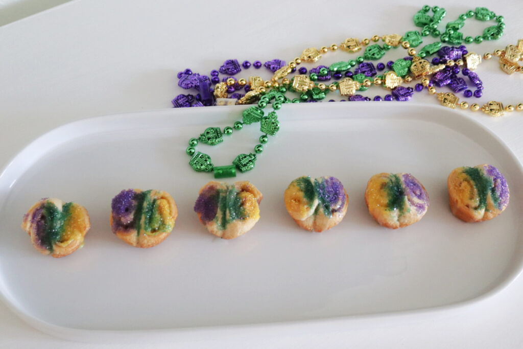 Six King cake bites on a white platter with Mardi Gras beads on the table.