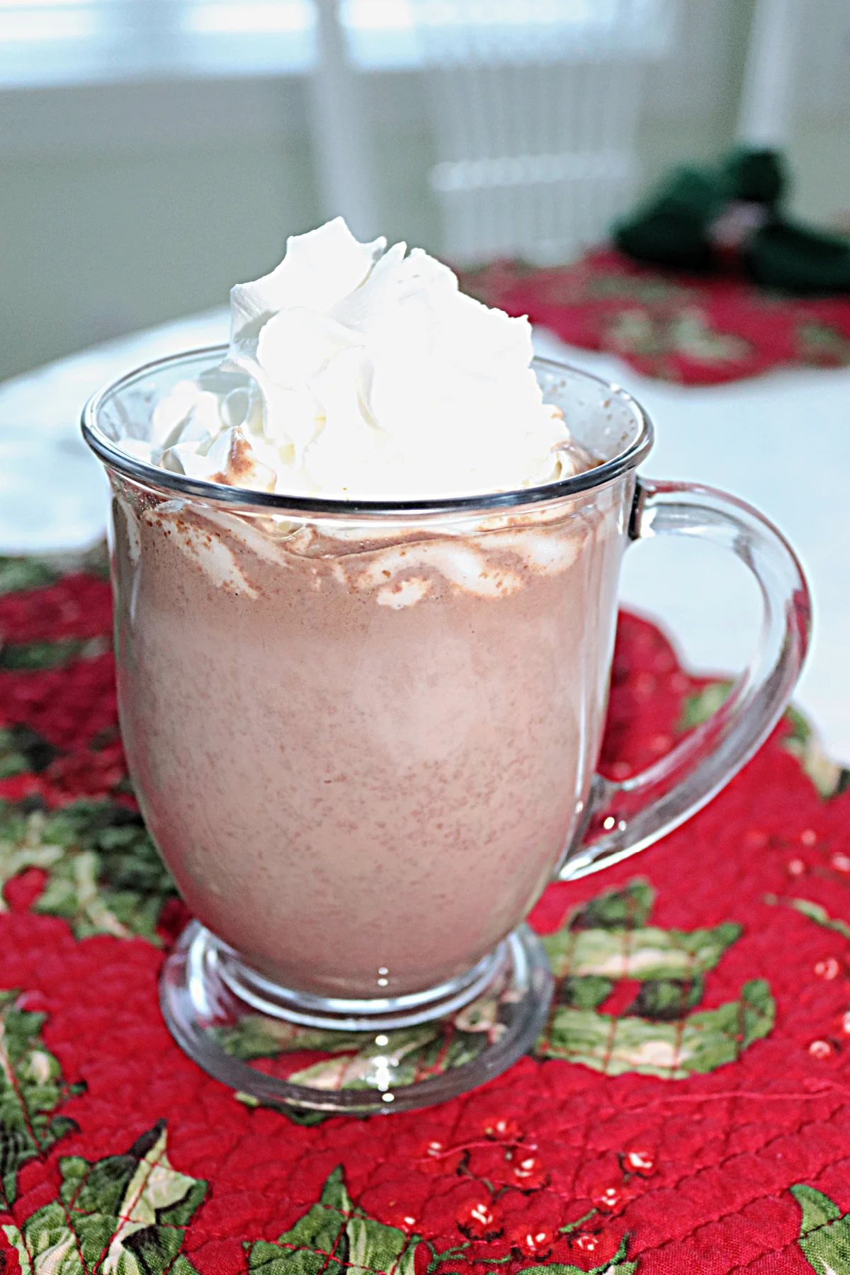 Ultimate hot chocolate in a glass mug with whipped cream on top sitting on a Christmas placemat.