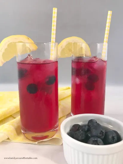 2 glasses of blueberry lemonade garnished with a lemon slice and a cup of blueberries on the counter.
