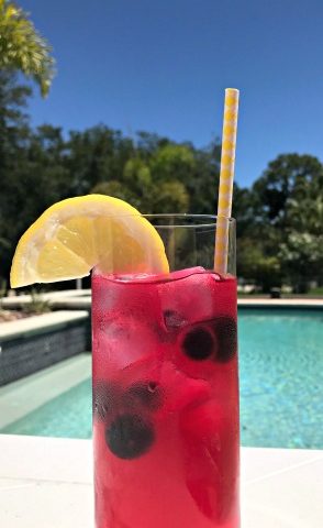 A glass of blueberry lemonade with a lemon slice garnish with a pool in the background.