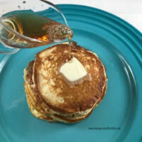 Buttermilk pancakes on a blue plate with a pat of butter on top and syrup being poured over them.