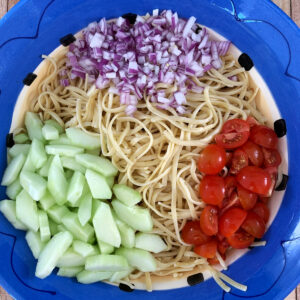 Overhead photo of pasta, red onion, cucumber and tomato in a colorful bowl ready to be mixed into linguine salad.