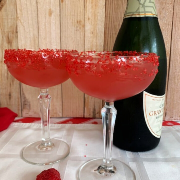 Two coupe glasses filled with a pink flirtini cocktail with a bottle fo sparkling wine in the background.