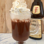 A clear mug of Frangelico hot chocolate with whipped cream and a bottle of Frangelico in the background.