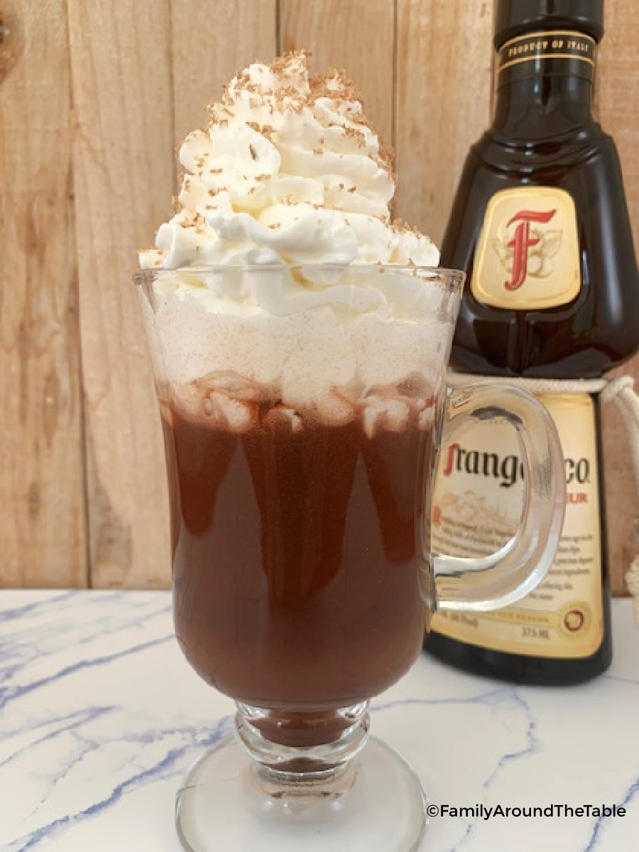 A clear mug of Frangelico hot chocolate with whipped cream and a bottle of Frangelico in the background.