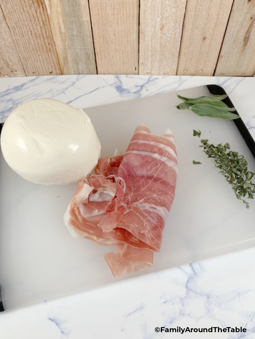 Mozzarella, prosciutto and chopped sage on a cutting board.