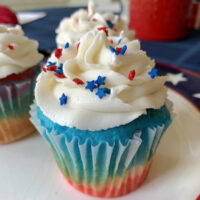 A red, white, and blue cupcake with white frosting and sprinkles on a plate.
