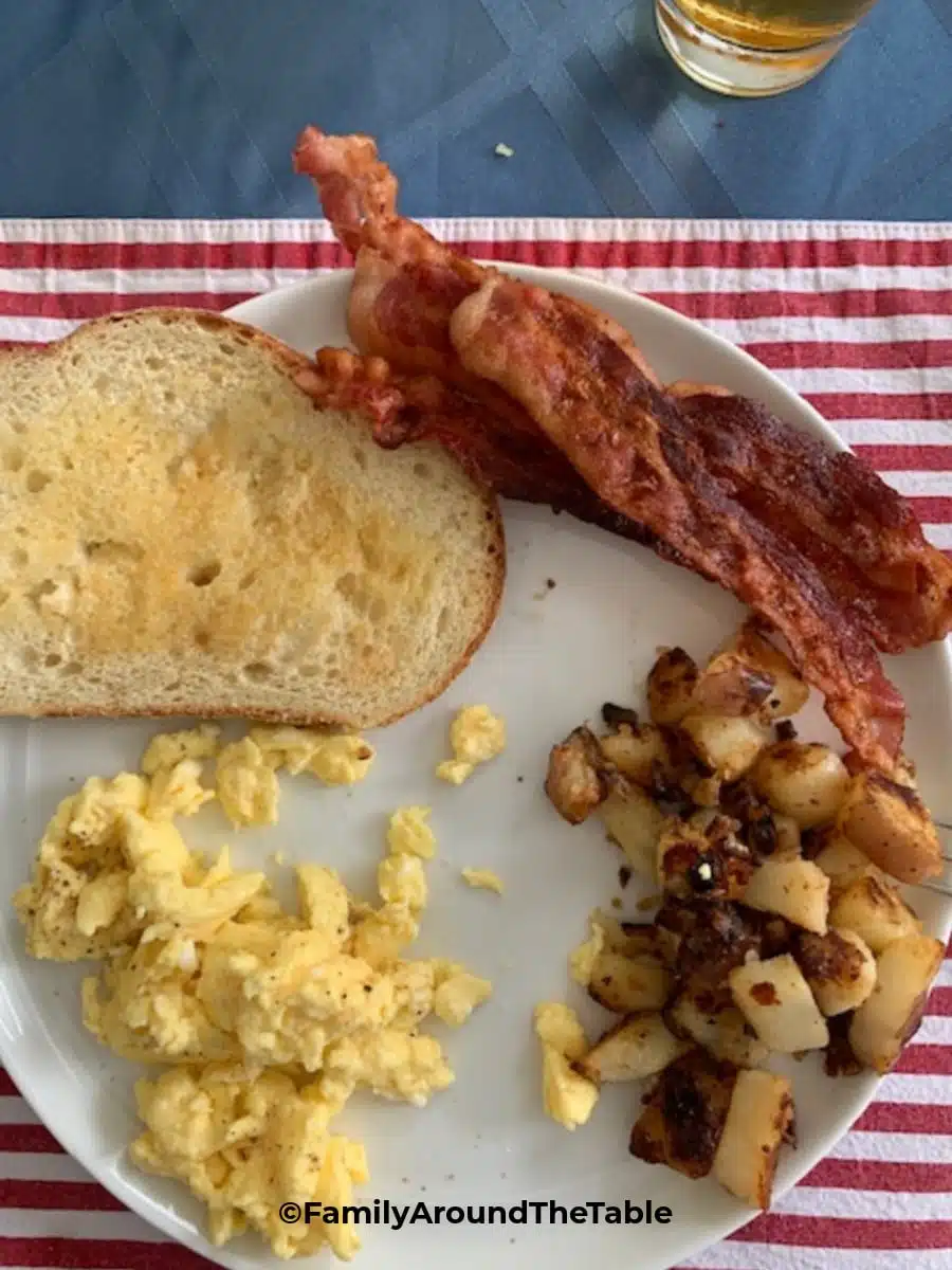 A breakfast plate with bacon, scrambled eggs, a piece of toast and diner-style breakfast potatoes and onions.