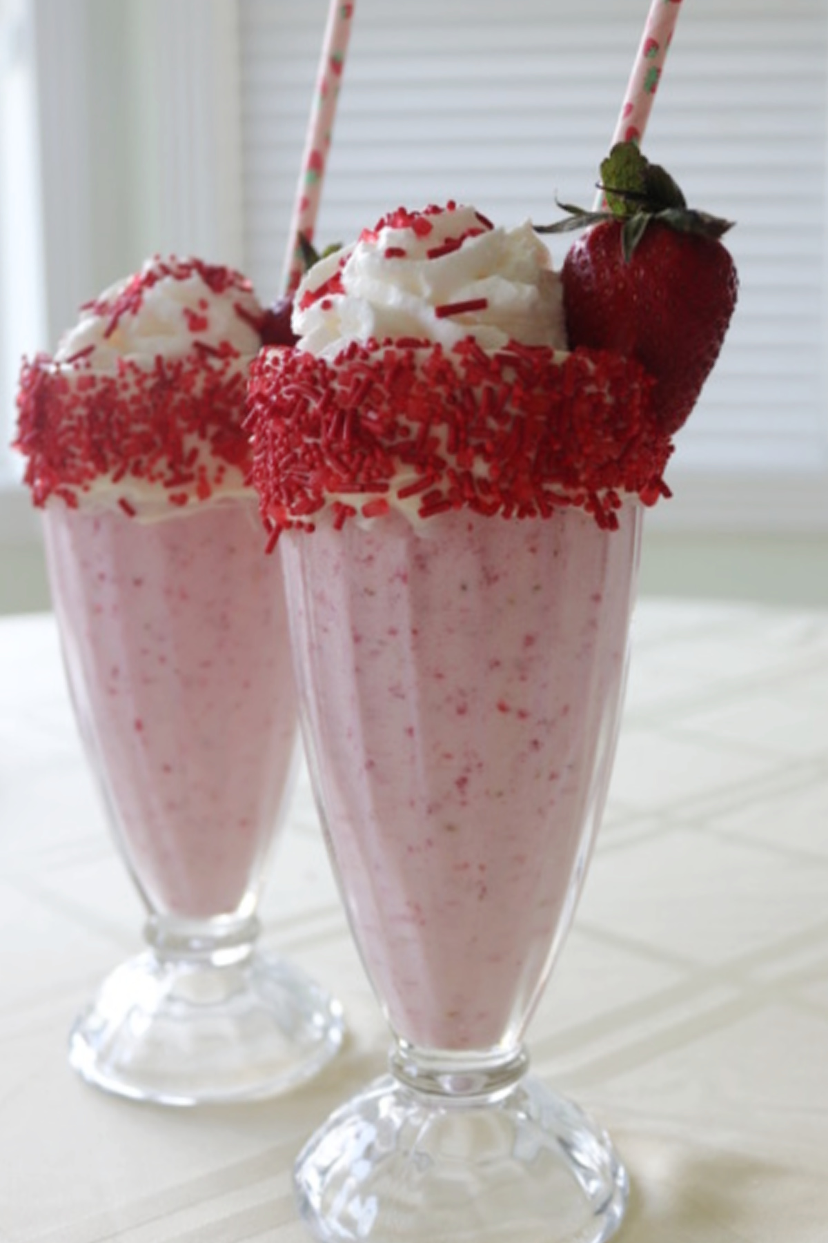 Two fresh strawberry milkshakes with whipped cream and sprinkles garnished with a strawberry.