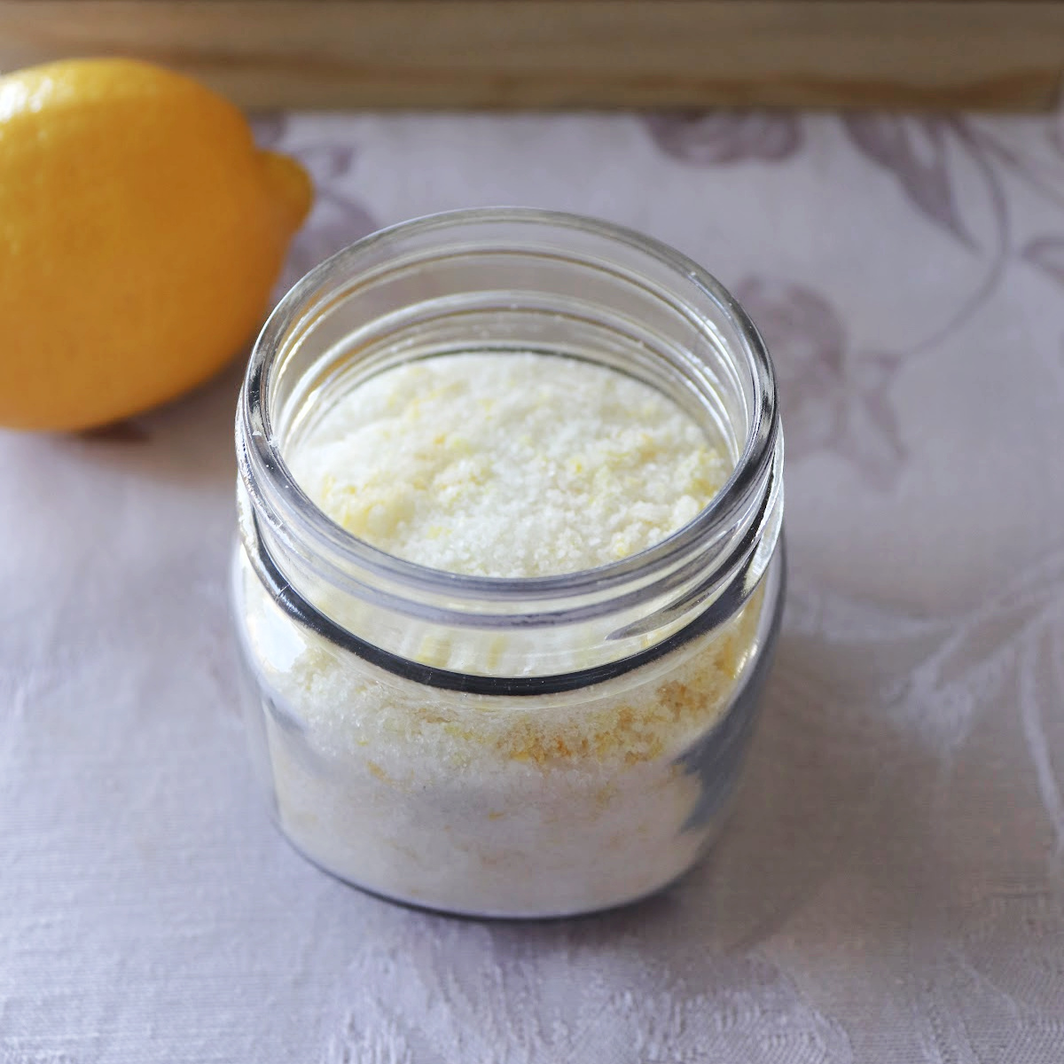 A glass jar with lemon salt sitting on a table with lemons around it.