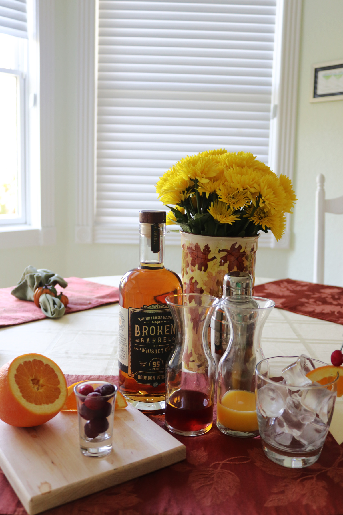 A bottle of bourbon, carafes with orange and cranberry juice, a glass with ice and half of an orange on a cutting board all on a table.