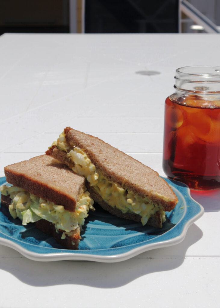 Southern egg salad sandwich on a blue plate with a glass of iced tea next to it, sitting on a white table.