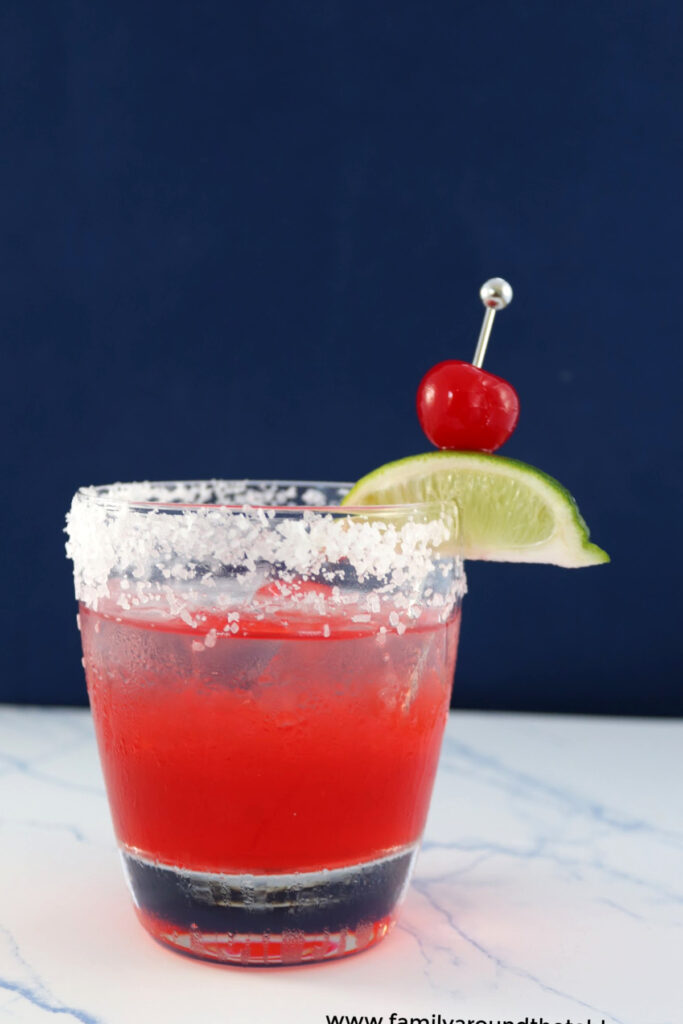 A glass with red liquid and a salted rim with a cherry lime garnish on a counter.