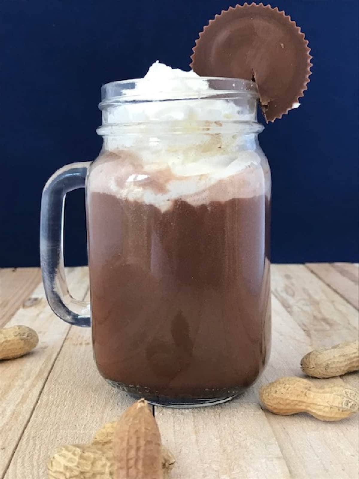 A glass mug of spiked peanut butter hot chocolate with whipped cream garnished with a Reese's peanut butter cup.