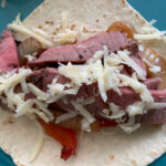 Steak, peppers, onions, and cheese on a flour tortilla to make grilled steak fajitas.