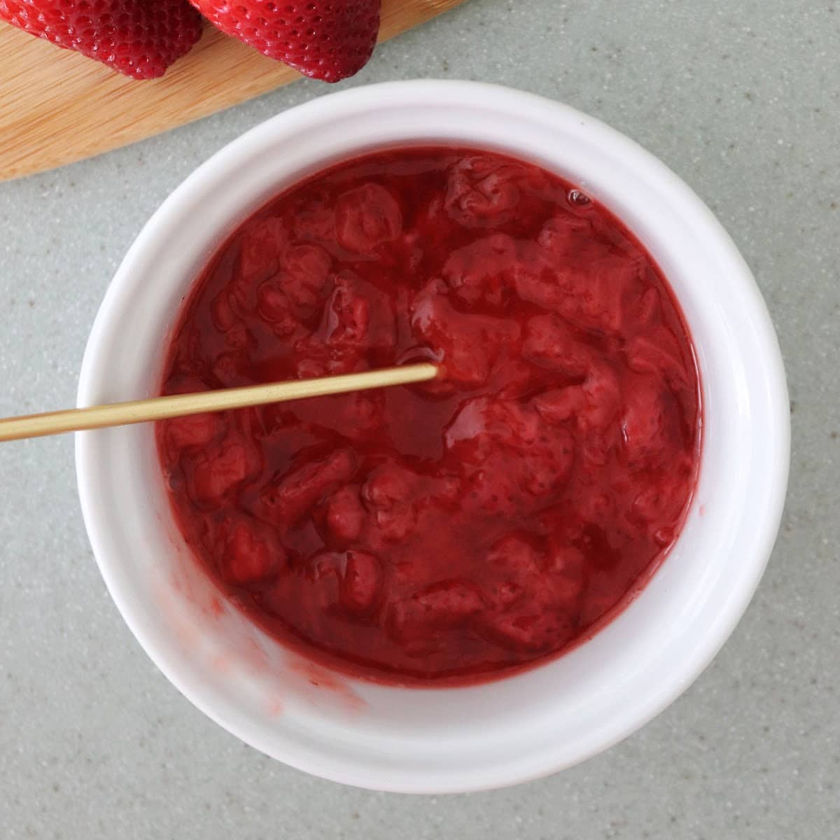 Overhead view of fresh strawberry sauce in a white ramekin with a gold spoon in it.