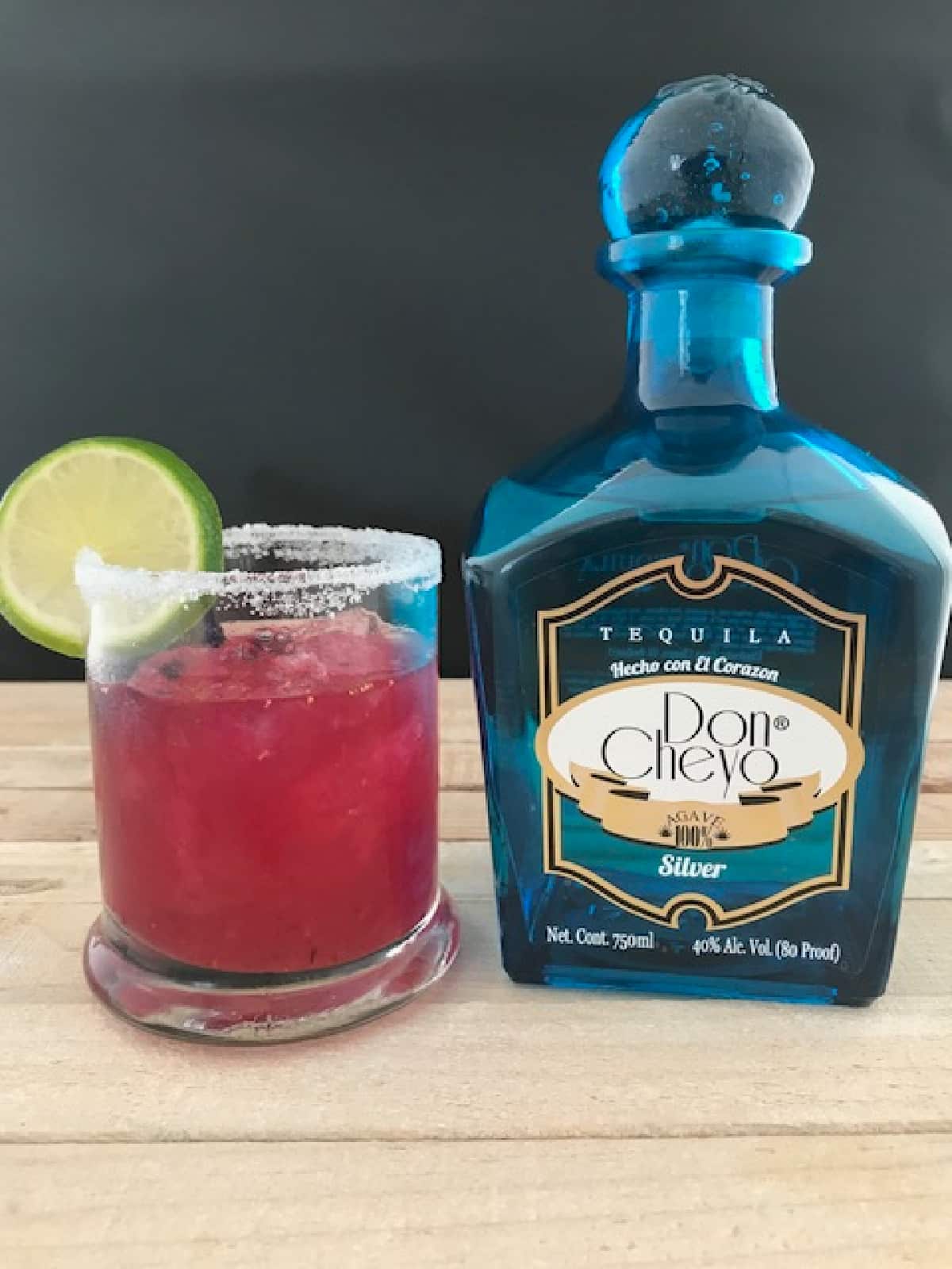 A blueberry margarita garnished with a lime wheel next to a blue bottle of tequila.