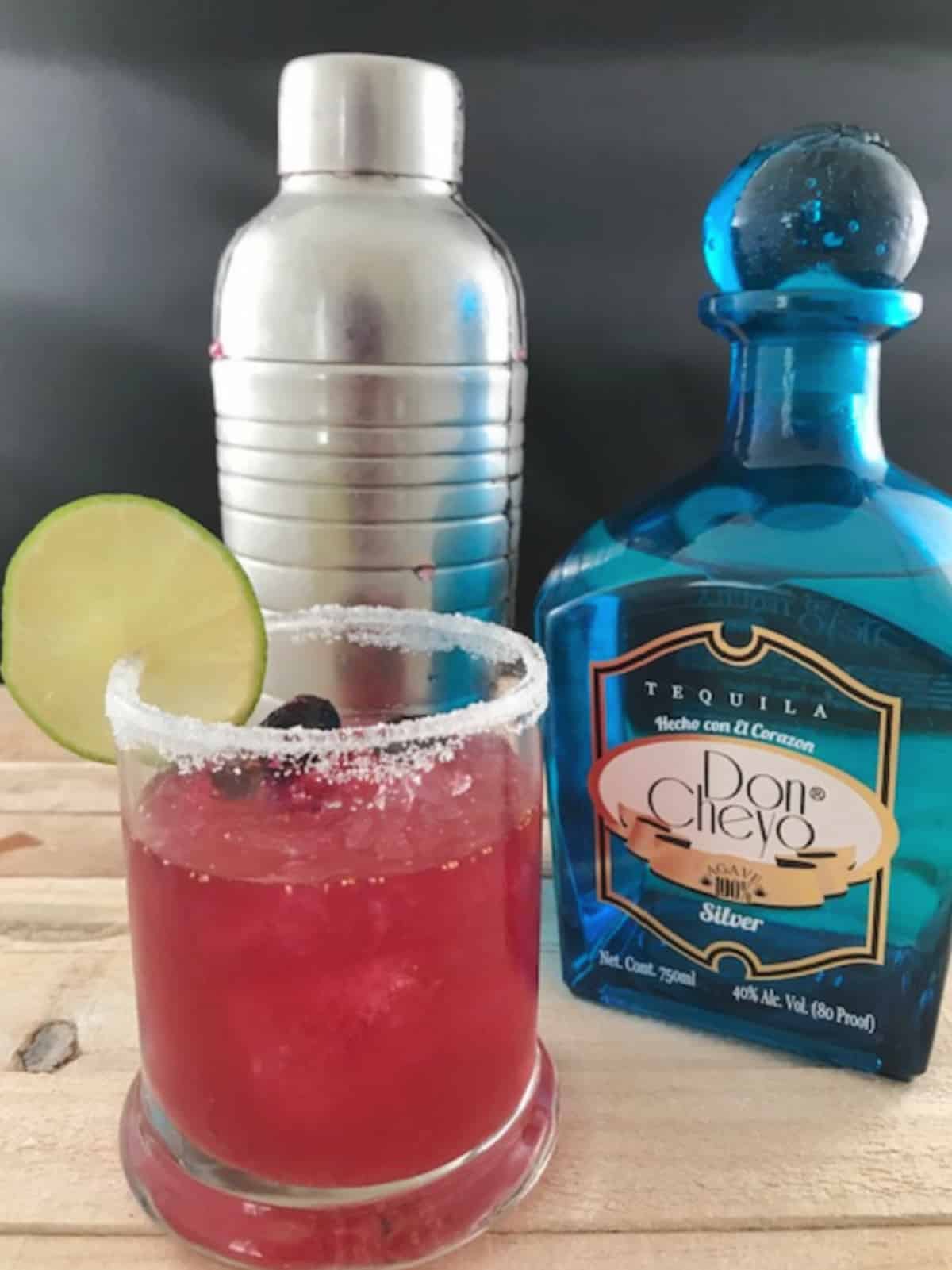 Blueberry margarita garnished with a lime wheel next to a silver cocktail shaker and blue tequila bottle.