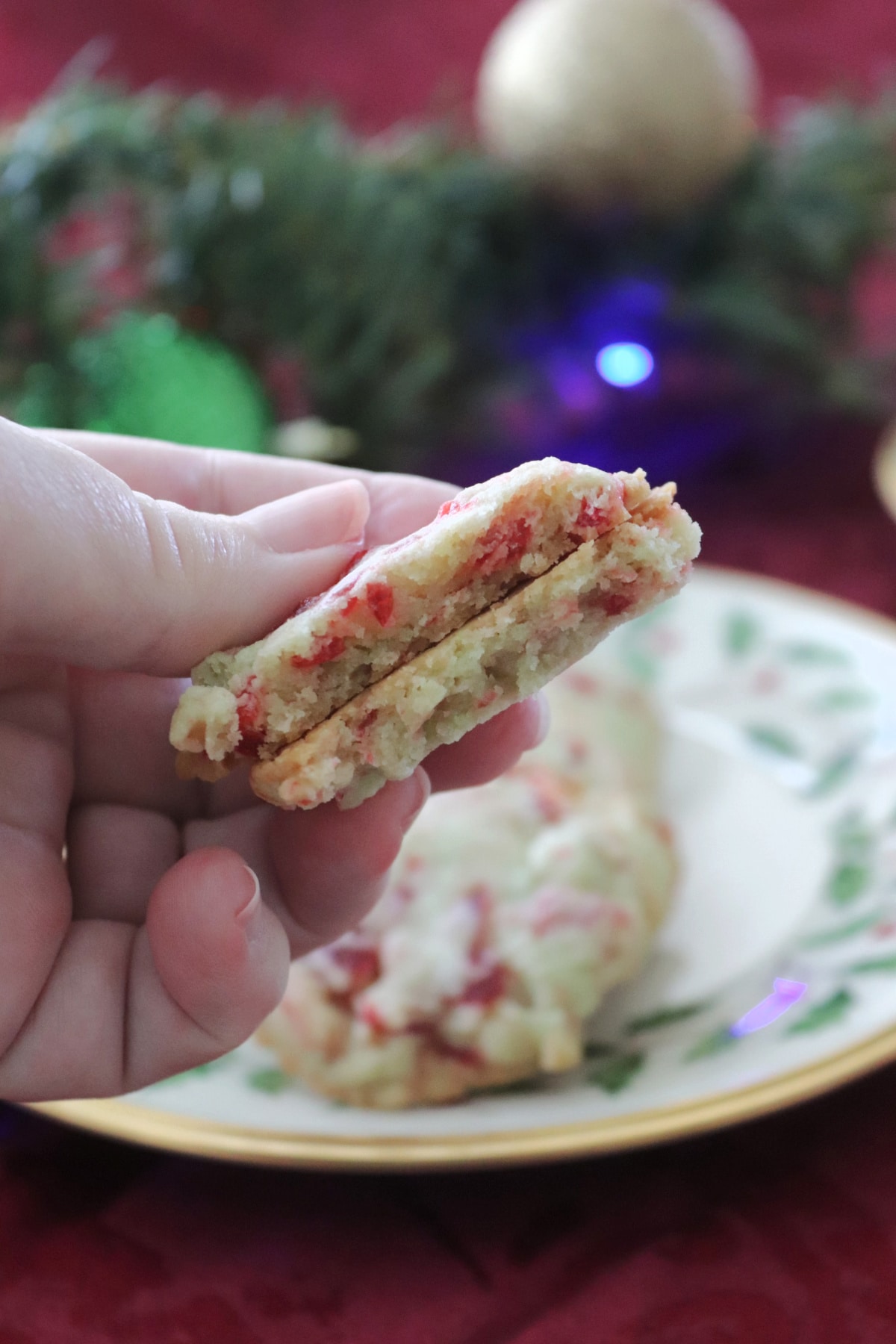 A cherry pistachio cookie that's been broken in half being held over a plate with more cookies on it.