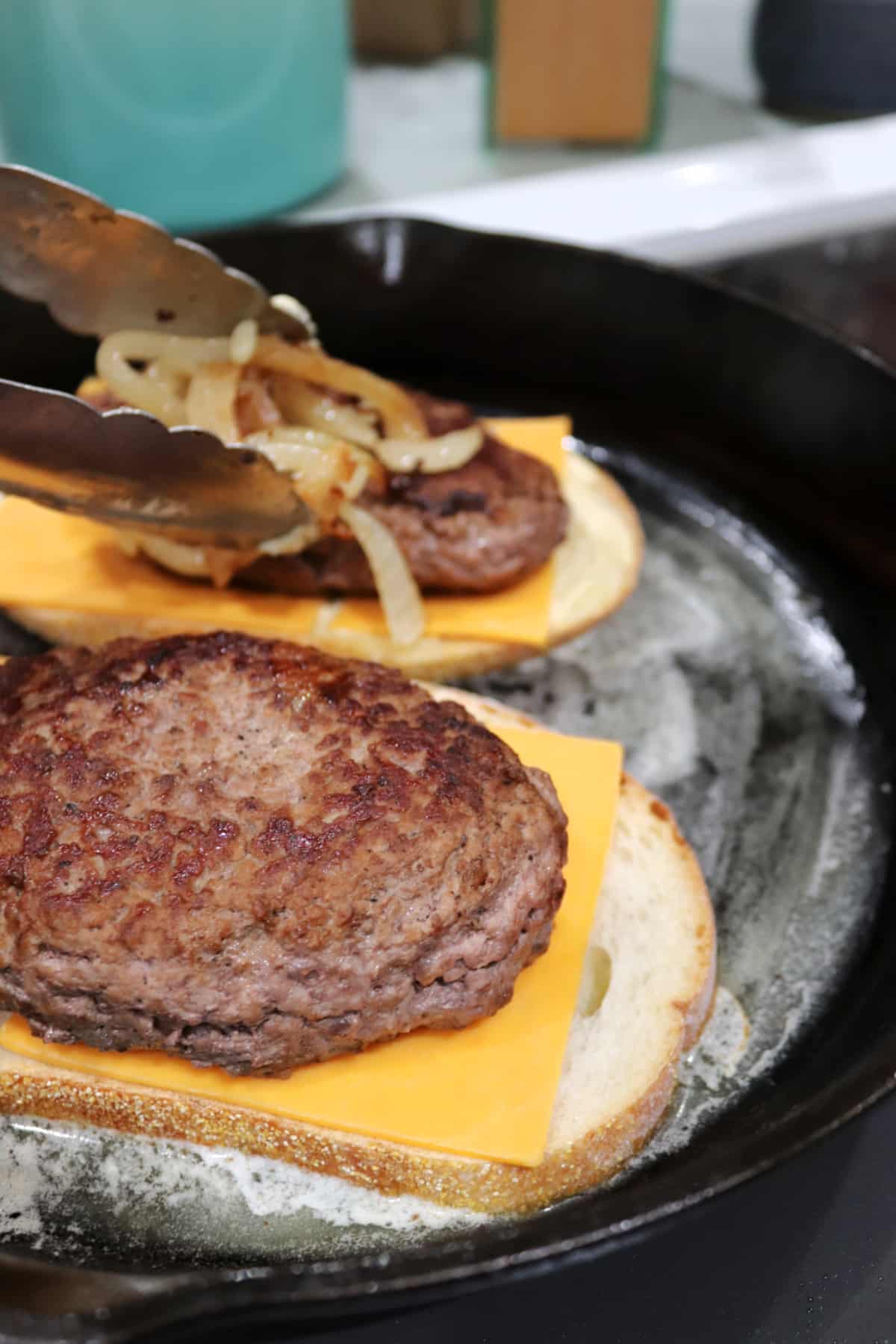 Two patty melt sandwiches in a skillet with caramelized onions being placed on one.