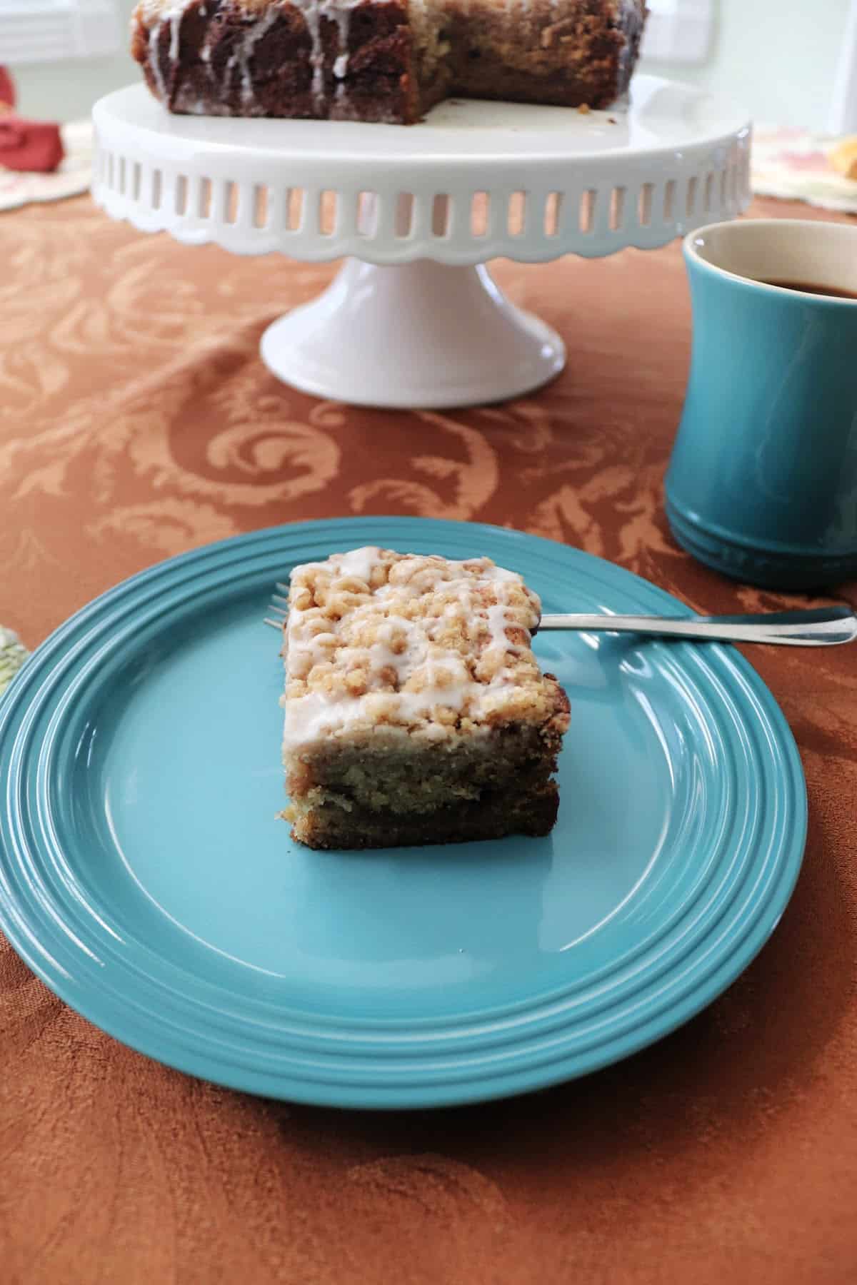 A slice of banana crumb cake on a blue plate with a cup of coffee behind it and a cake plate with the cake on it behind it.