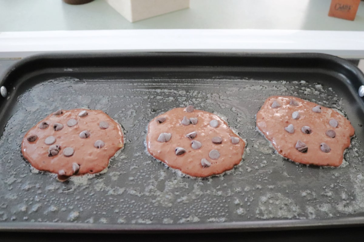 Three chocolate chocolate chip pancakes on a flat griddle.