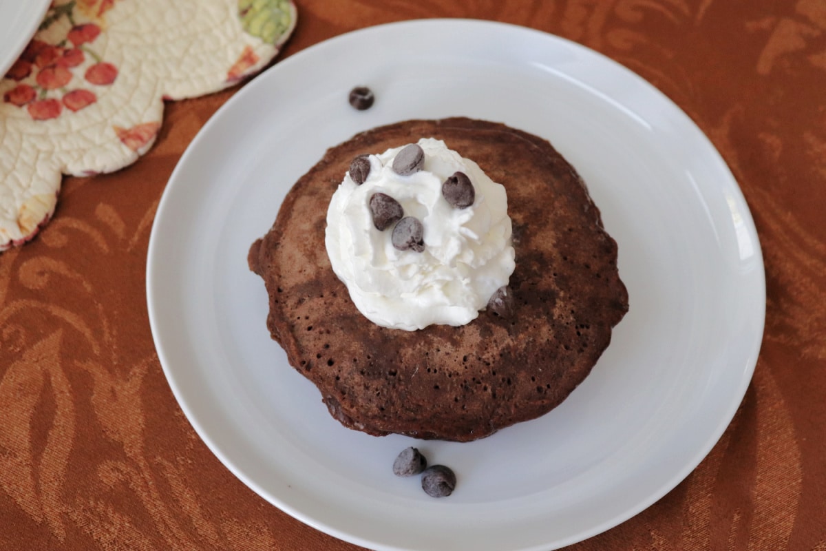 Overhead view of chocolate chocolate chip pancakes on a white plate garnished with whipped cream and chocolate chips.