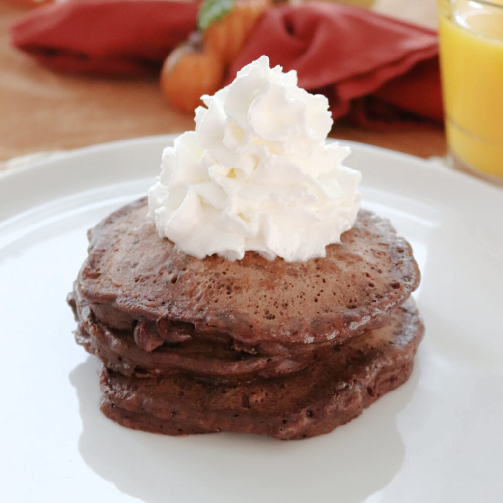 Chocolate chocolate chip pancakes stacked on a white plate topped with whipped cream.